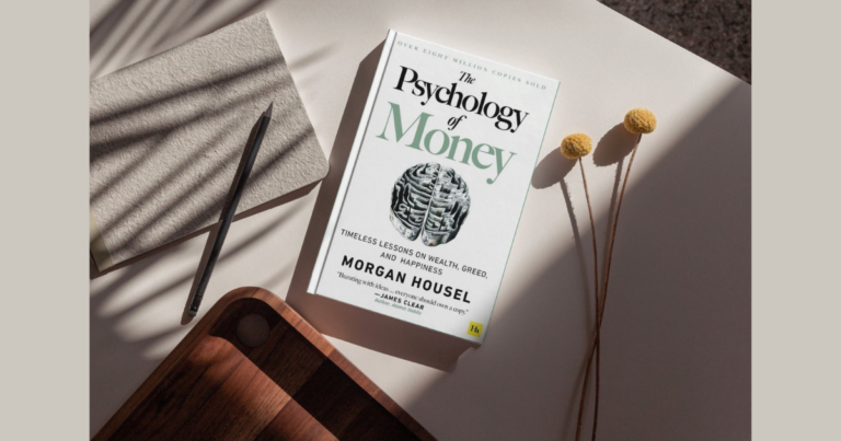 The Psychology of Money by Morgan Housel book with notebook, pencil, and dried flowers on a neutral desk