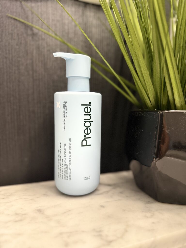 Prequel Urea Advanced Relief Moisturizing Milk bottle displayed on a bathroom countertop next to a plant.